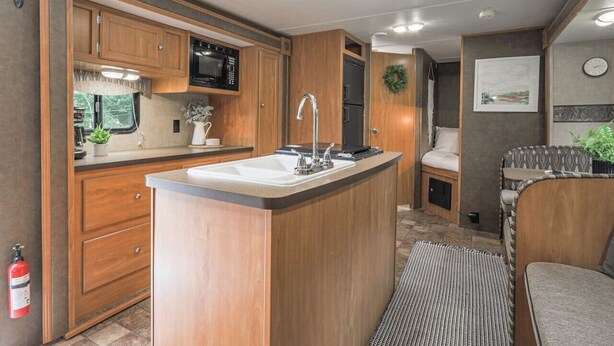 A compact RV kitchen with wooden cabinets, a central island with a sink, a microwave, and a refrigerator.