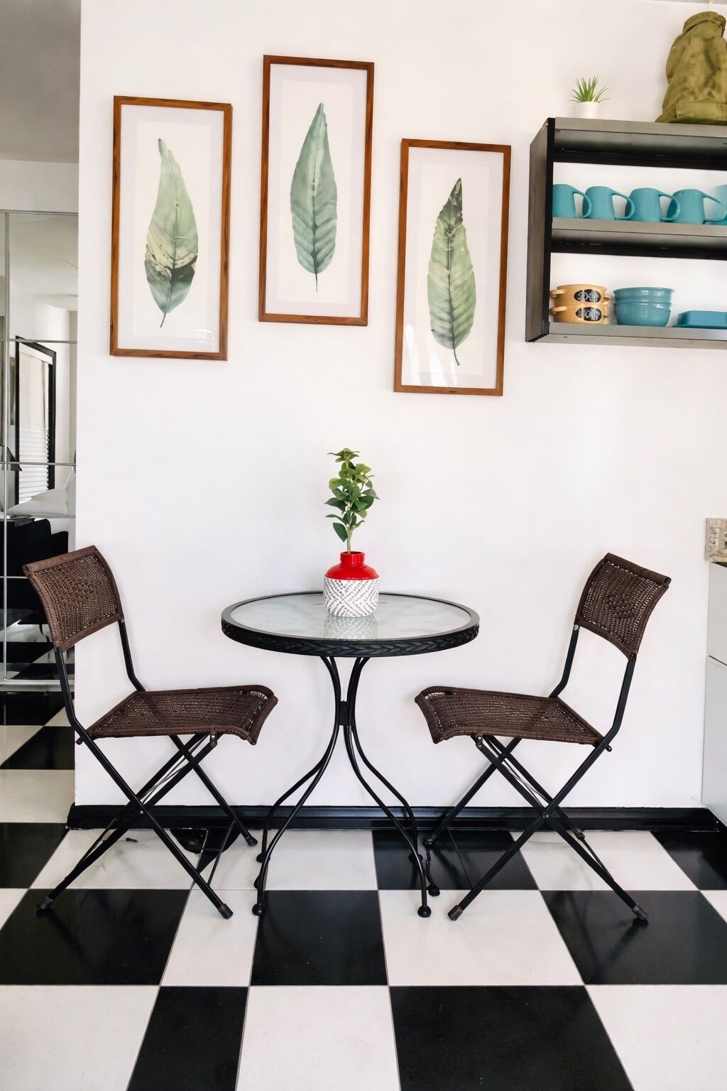Un coin repas avec un sol à carreaux, deux chaises en osier, une table ronde ornée d’un vase rouge et trois œuvres d’art représentant des feuilles, encadrées et accrochées au mur.
