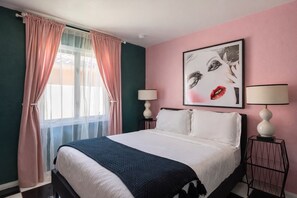 A hotel room with a bed, two bedside lamps, a window with pink curtains, and a wall art piece. - The Rosemary House La Quinta No.218742 (La Quinta)