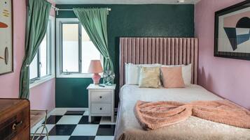 A bedroom with a checkered floor, a bed with a pink blanket, a bedside table with a lamp and fan, and a dresser.