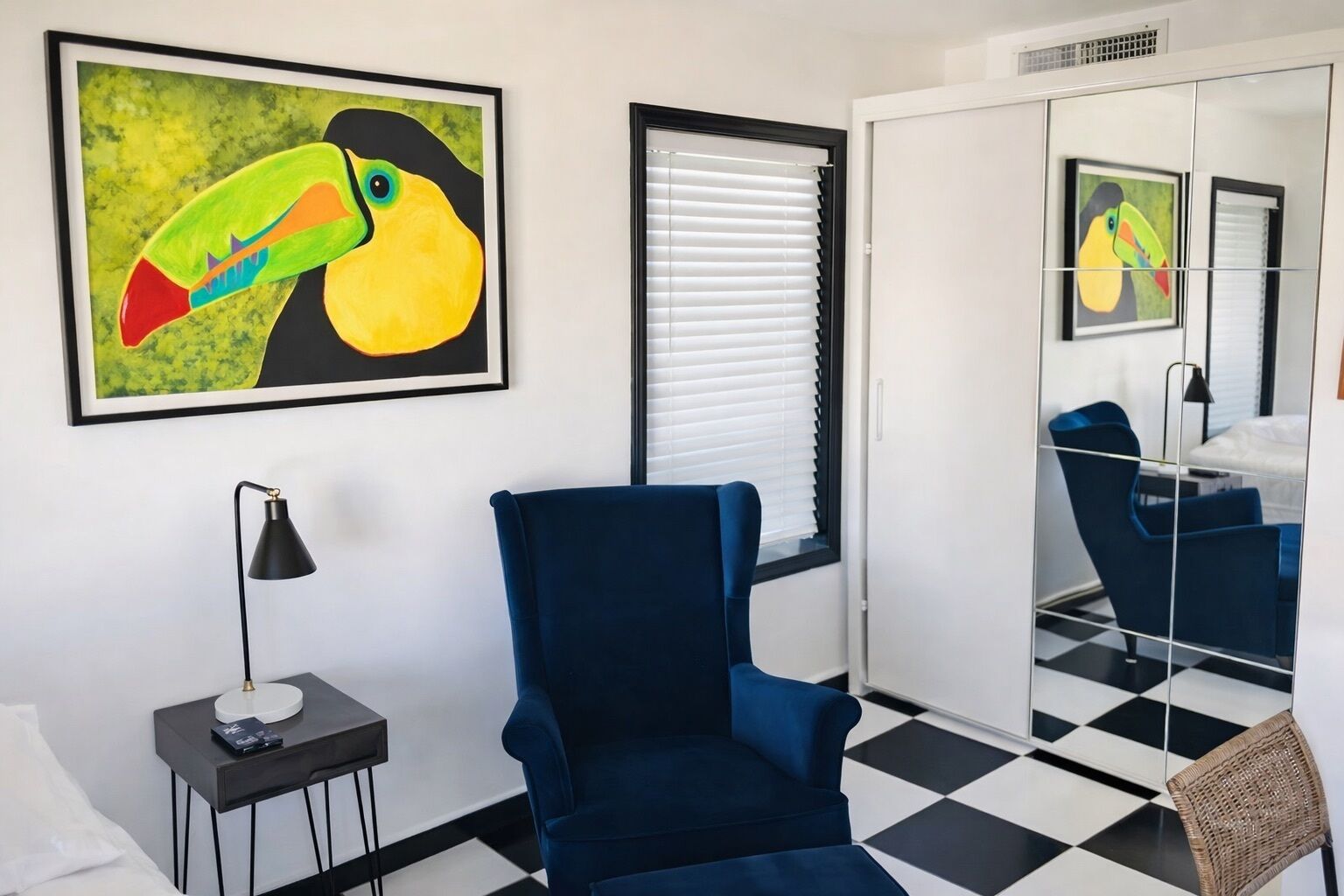 A room with a blue armchair, a small table with a lamp, a checkered floor, and a large mirror reflecting the room.
