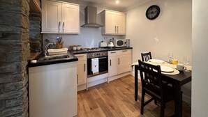 A compact kitchen with white cabinets, a stove, and a sink. There is a dining table with chairs and a clock on the wall. - The Cosy Cottage Retreat! Long Stay Discounts (Llantrisant)
