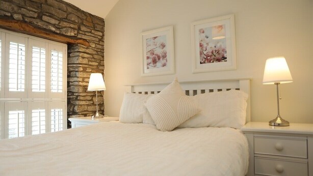 A bedroom with a stone wall, a bed with white bedding, two bedside lamps, and two framed floral pictures on the wall. - The Cosy Cottage Retreat! Long Stay Discounts (Llantrisant)