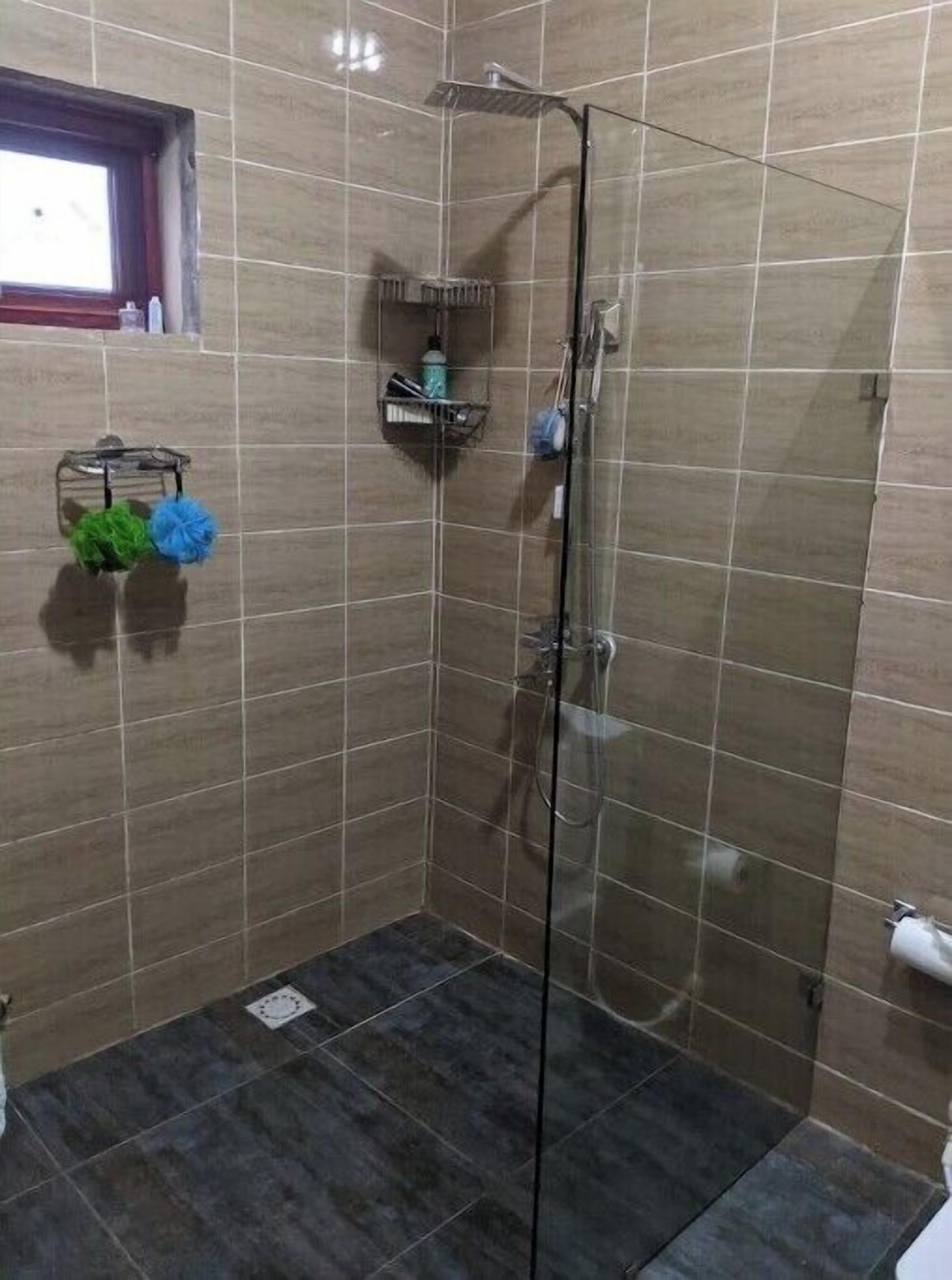 Et bruseområde med glasdør, flisebeklædte vægge, et lille vindue og en hylde med toiletartikler.