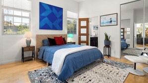 Une chambre moderne avec un grand lit, une œuvre d’art géométrique bleue, une porte en bois et une vue sur l’extérieur.