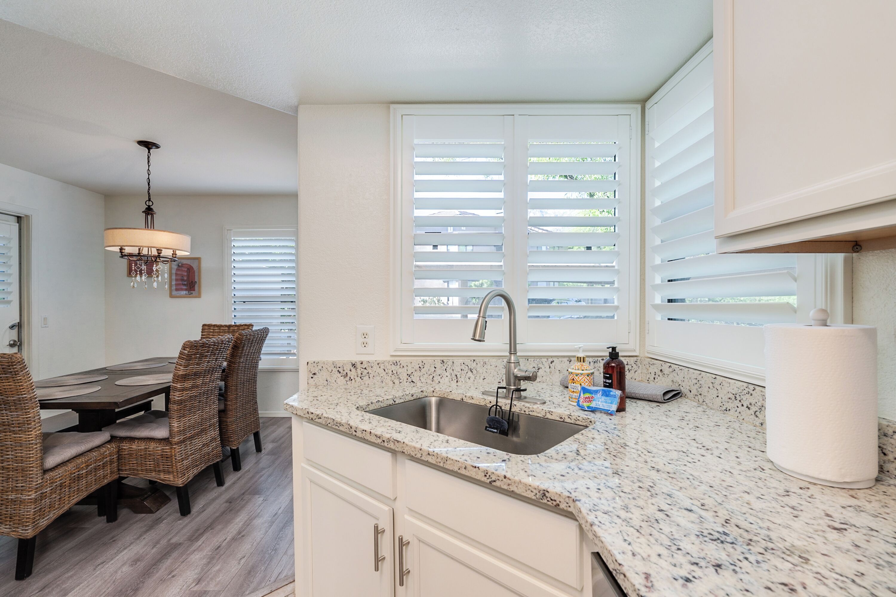 A kitchen with a granite countertop, white cabinets, a stainless steel sink, and a dining area with wicker chairs.