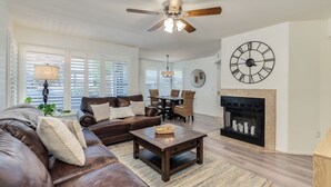 A spacious living room with a brown leather sofa, a wooden coffee table, a fireplace, and a large wall clock.
