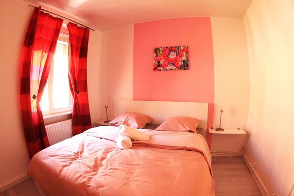 A bedroom with a bed, red curtains, a nightstand, a lamp, and a painting on the wall.