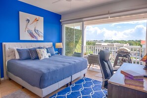 A bedroom with a large bed, a desk with books, a chair, and a balcony with a view of the sky and greenery. - Relaxing Waterfront Getaway in Puerto Aventuras (Puerto Aventuras)