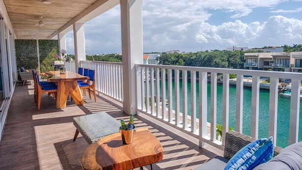 A balcony with a wooden table, blue chairs, and a view of a body of water. - Relaxing Waterfront Getaway in Puerto Aventuras (Puerto Aventuras)