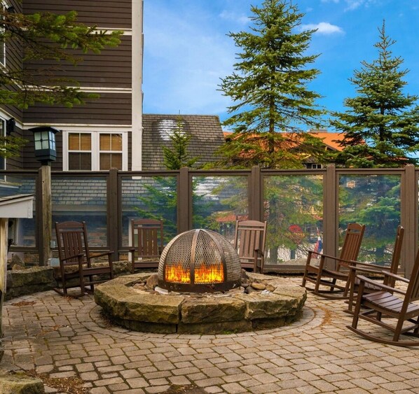 A circular stone fire pit with chairs around it, set on a paved patio with a view of trees and a building in the background.