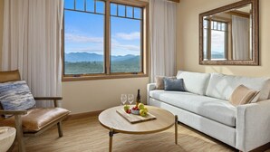 Une pièce comprenant un canapé, un fauteuil, une table ronde avec des fruits et des verres, une grande fenêtre donnant sur la montagne et un miroir au mur.