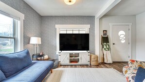 A modern living room with a large flat-screen TV, a blue sofa, a patterned armchair, and a white entertainment unit.