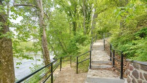 A concrete staircase with metal railings leading up a stone wall, surrounded by dense green foliage and a body of water in the background. - Riverfront Shenandoah Getaway w/ Kayaks & Tubes! (Shenandoah)