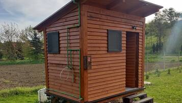 A small wooden cabin with a porch and two windows, situated in a grassy area with trees in the background.