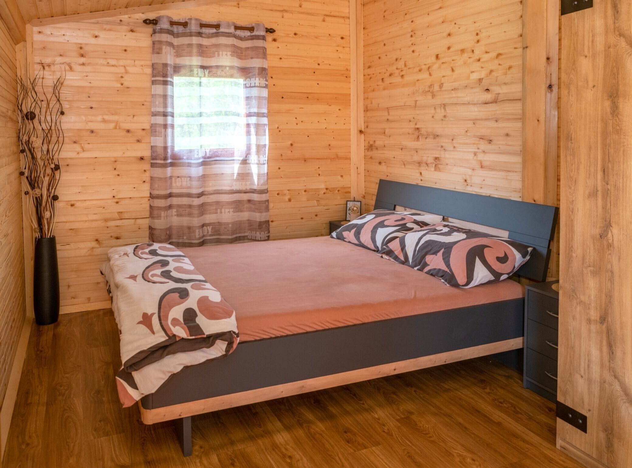 Une chambre à coucher dans une cabane en bois, avec un lit, une table de chevet, une fenêtre avec des rideaux et un vase décoré de branches.