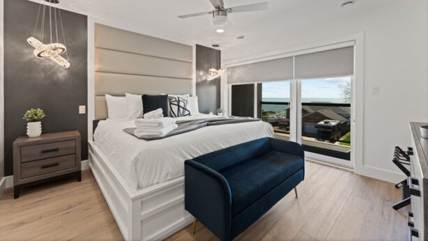 A modern bedroom with a large bed, a dark blue bench, a ceiling fan, and a view of the ocean through a sliding glass door. - Luxury 171-7 Condos with Magnificent Lakeviews (Leamington)