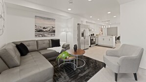 A modern living room with a grey sofa, a round coffee table, and a grey armchair. A kitchen is visible in the background with white cabinets and stainless steel appliances. - Luxury 171-7 Condos with Magnificent Lakeviews (Leamington)