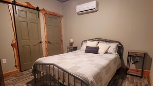 Un dormitorio con estructura de cama metálica, colcha acolchada, mesita de noche con lámpara y suelo de madera.