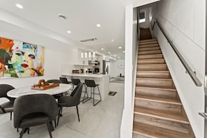 Una cocina moderna con armarios blancos y una isla central, un comedor con una mesa redonda y sillas, y una escalera de madera.