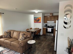 A modern living room with a brown leather sofa, a dining table with chairs, and a kitchen area with wooden cabinets and a stove. - Buffalo Cabin - Near T.R.N.P. (Belfield)