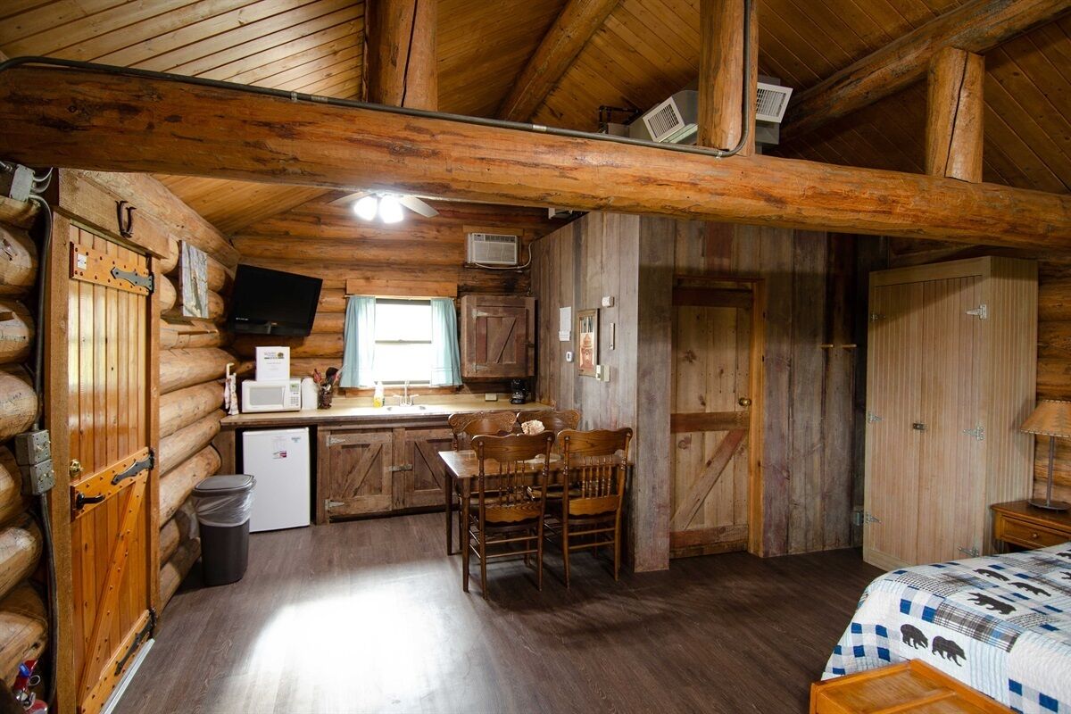 A log cabin interior with a kitchen area, wooden dining table, and a bed with a patterned bedspread.