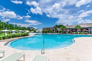 A large outdoor swimming pool with lounge chairs and umbrellas, surrounded by greenery and a building with an American flag.