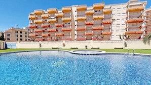 En swimmingpool med en klar blå vandoverflade, omgivet af en lav mur. I baggrunden ses en flereetagers bygning med altaner.