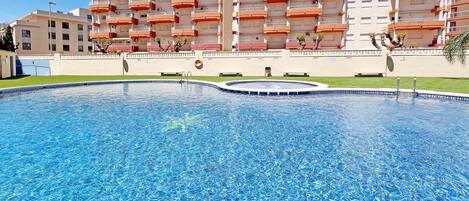 Una piscina con superficie de agua azul cristalina, rodeada por un muro bajo. Al fondo, un edificio de varios pisos con balcones.