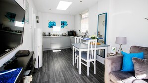 Une cuisine moderne avec des meubles blancs, une table à manger et des chaises. On y trouve un téléviseur posé sur un meuble, une machine à café et un four à micro-ondes.