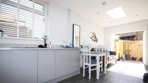 Une cuisine moderne avec des meubles blancs, un coin repas comprenant une table et des chaises blanches, et une grande fenêtre avec des stores.