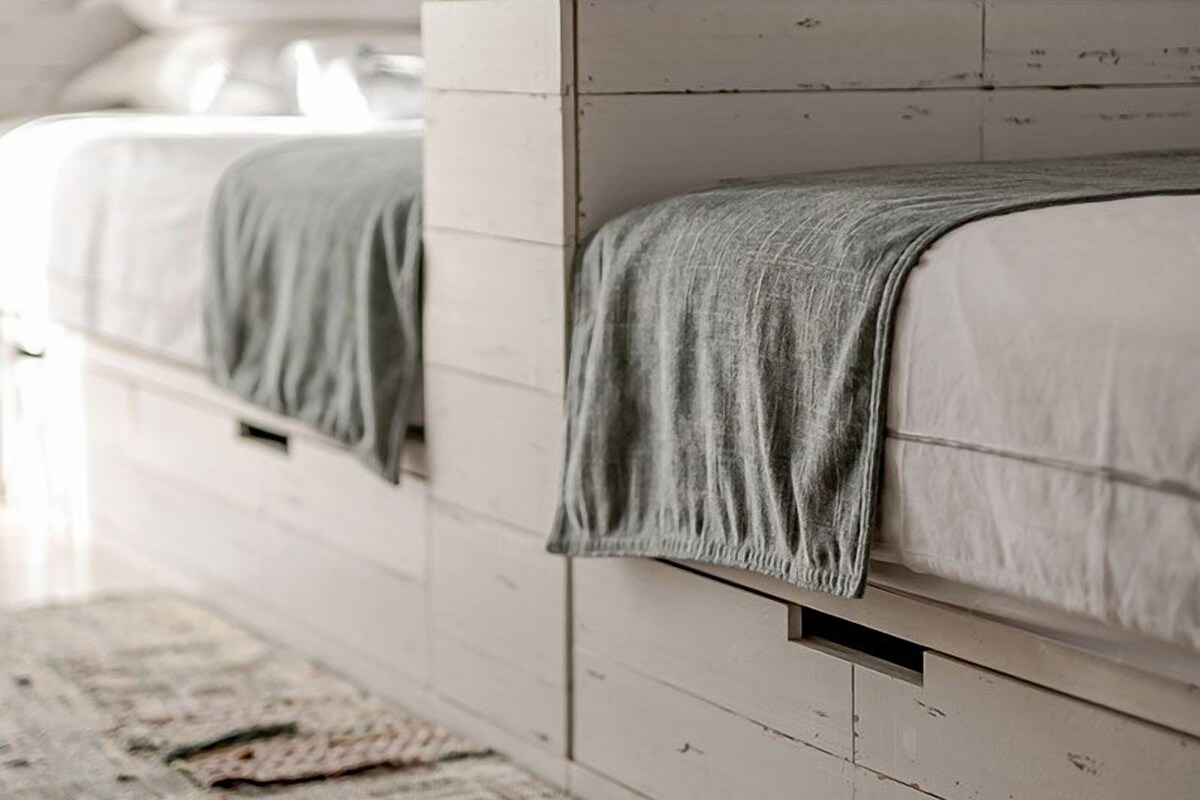 A bedroom with two beds, a wooden headboard, a white wall, and a small shelf above the beds.