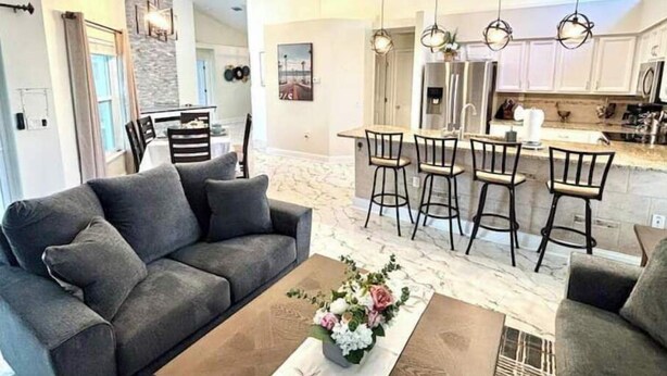 A modern living room with a grey sofa, a coffee table with a floral centerpiece, and a dining area with bar stools and pendant lighting. - Luxurious Relaxation Home - Pool Games / 8PPL-4BR (Tampa)