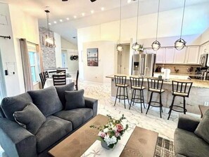 Relax in this inviting living space with a sofa, armchair, and coffee table, opening to a kitchen with a central island and bar stools.