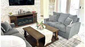 A living room with a grey sofa, a wooden coffee table, and a TV mounted on a stone wall. - Luxurious Relaxation Home - Pool Games / 8PPL-4BR (Tampa)