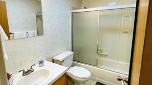 A bathroom with a white sink, mirror, and toilet, a shower with a glass door, and a light fixture on the ceiling. - Waterfront unit! Walk to shops, dining, & trails! (Anchorage)