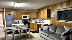 A compact kitchen with wooden cabinets, a refrigerator, a microwave, and a dining area with a table and chairs. - Waterfront unit! Walk to shops, dining, & trails! (Anchorage)