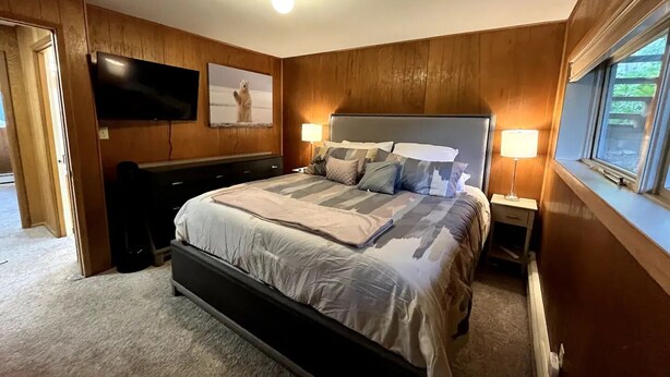 A bedroom with a bed, bedside lamps, a TV mounted on the wall, and a window. - Waterfront unit! Walk to shops, dining, & trails! (Anchorage)