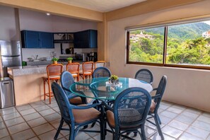 Une cuisine moderne avec des meubles bleus, un coin repas avec une table en verre et des chaises en osier, et une vue sur un paysage verdoyant par une grande fenêtre.