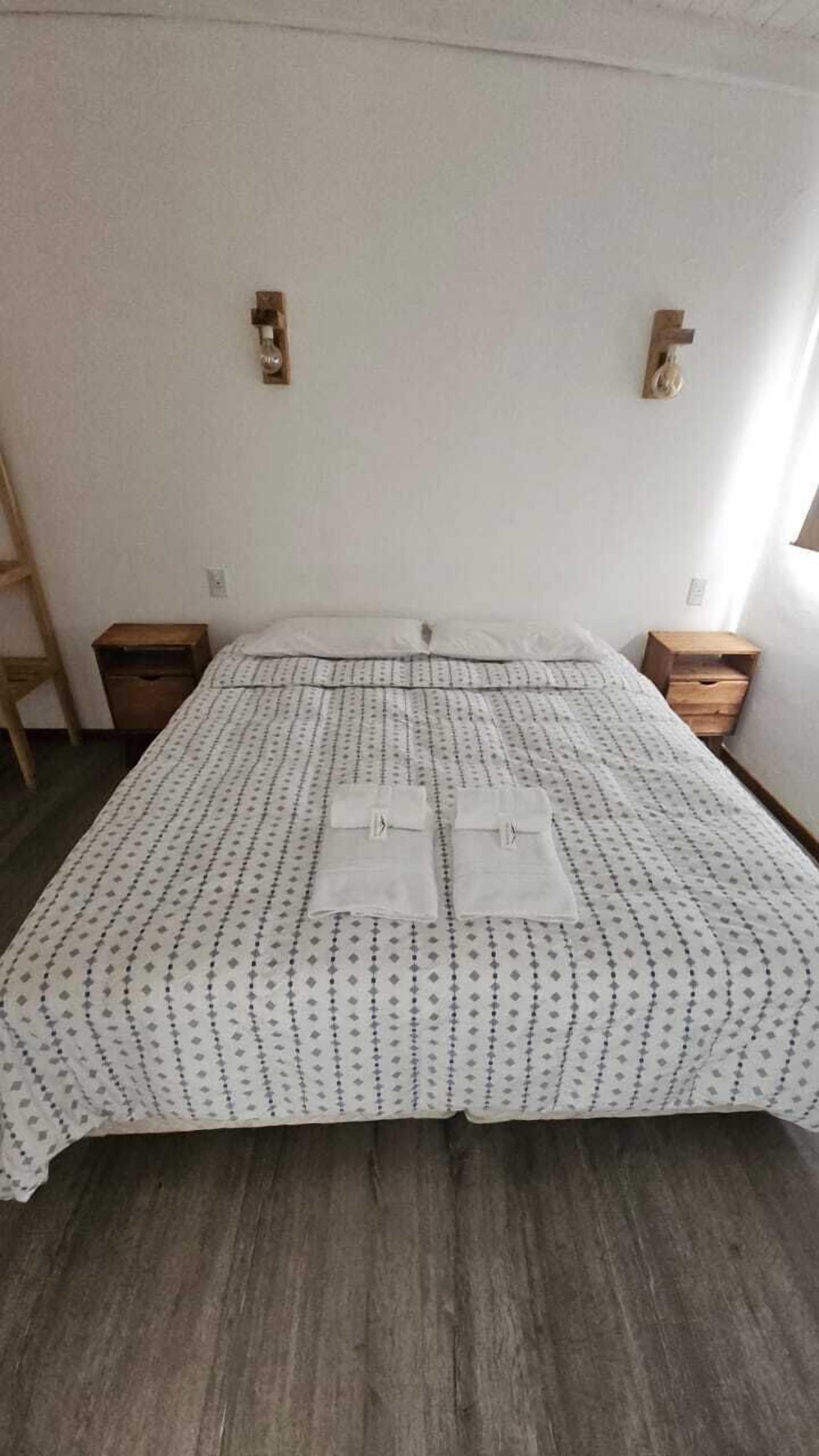 Un letto rifatto con cura, con una coperta a fantasia e due asciugamani piegati sopra. Ci sono due comodini in legno, uno per lato del letto.