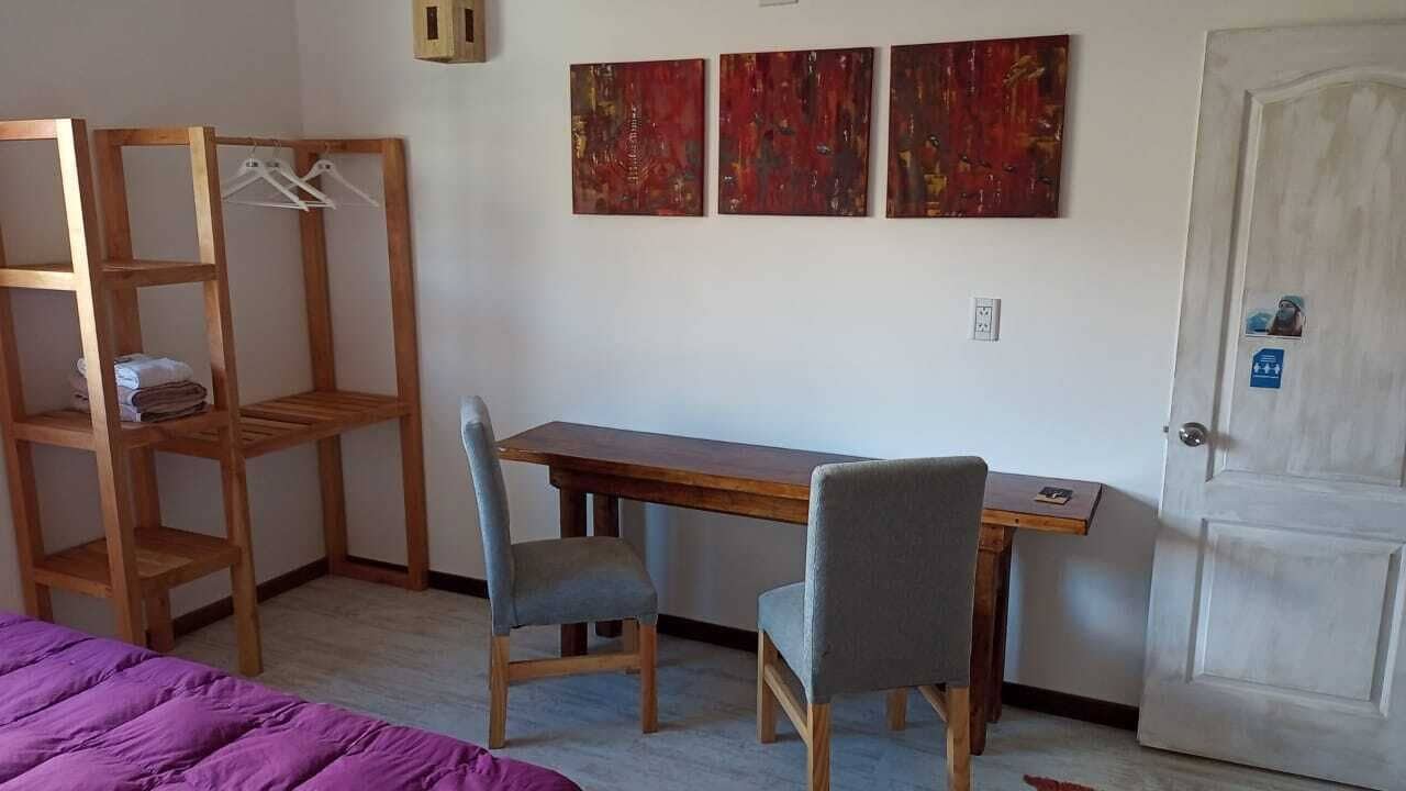 A room with a wooden desk, two chairs, a bed with a purple blanket, and three abstract paintings on the wall.