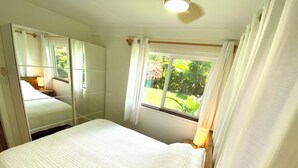 Een slaapkamer met een groot bed, een spiegel, een raam met gordijnen en uitzicht op groen.