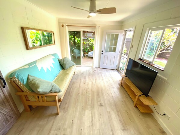 A room with a wooden floor, a sofa with cushions, a television on a wooden stand, and a door leading outside. - North Shore Country Getaway- steps to the ocean (Waialua)