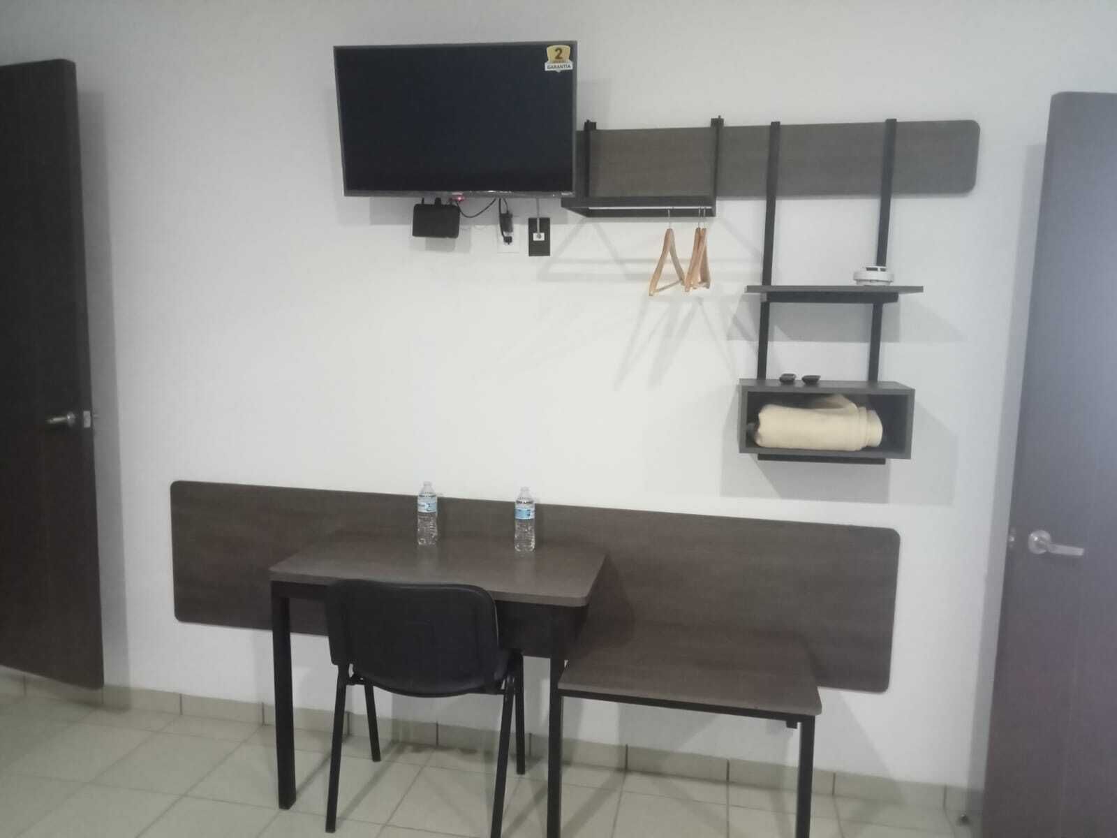 Una habitación pequeña con una televisión montada en la pared, un banco, una mesa con dos sillas y un estante con una toalla y una botella.
