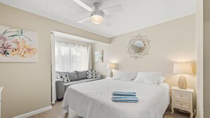 Ein Schlafzimmer mit einem großen Bett, einem Deckenventilator, einem Fenster mit Vorhängen und einem Nachttisch mit Lampe.