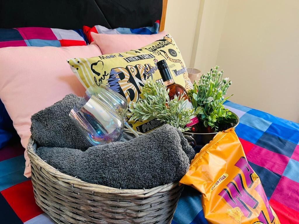 A basket with a bottle, a plant, and a gluten-free snack bag on a bed with a checkered blanket.