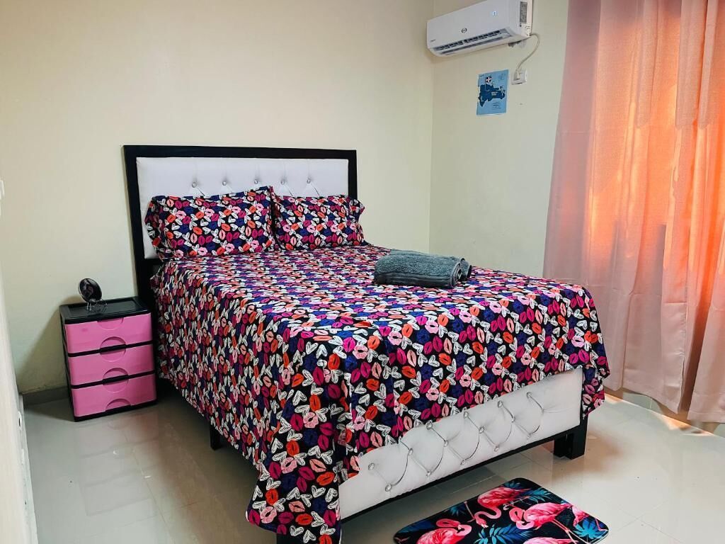A neatly made bed with a floral bedspread, a pink bedside table with drawers, and a wall-mounted air conditioner.