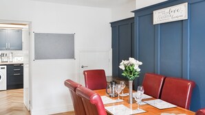 Un comedor con una mesa de madera dispuesta para cuatro personas, sillas tapizadas en rojo, un florero con flores blancas y un letrero en la pared.