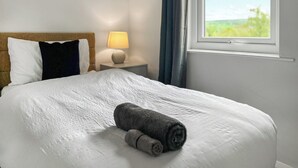 Una cama bien hecha con una toalla enrollada, una lámpara de noche y una ventana con vistas a zonas verdes.