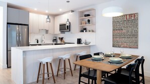 Eine moderne Küche mit weißen Schränken, einem Edelstahlkühlschrank, einer Mikrowelle und einer Kaffeemaschine. Ein Essbereich mit Holztisch und schwarzen Stühlen ist ebenfalls vorhanden.